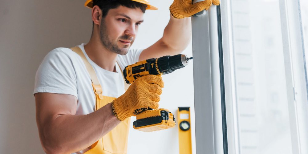 Handyman in yellow uniform installs new window by using automatic screwdriver. House renovation conception.