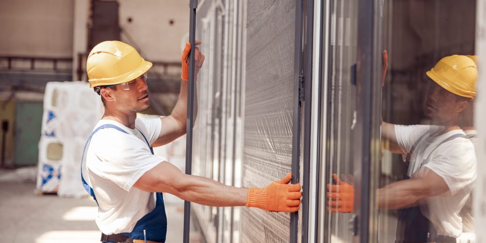 Handsome young man construction worker wearing safety helmet and work gloves while holding glass door
