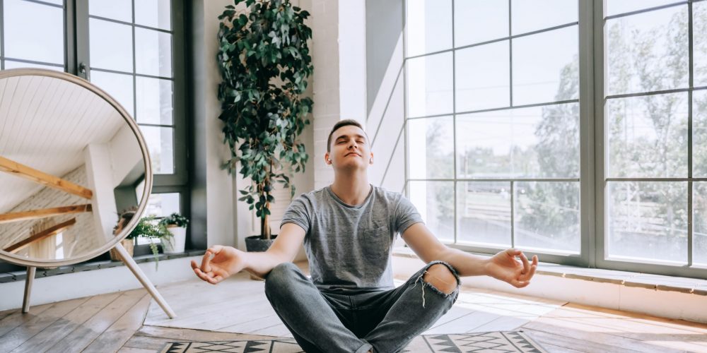 Pleased caucasian relaxed man in headphones sits crossed legs on floor near big window, tries to have break after work. Yoga and meditation concept.